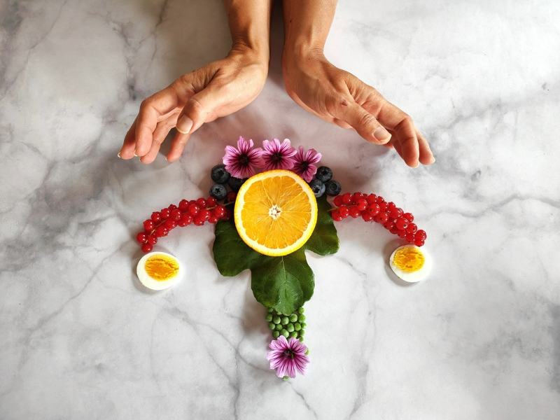 Kunstvolle Darstellung einer Gebärmutter aus Lebensmitteln: Orange, Beeren, Eier, Blumen und Erbsen, umgeben von schützenden Händen.